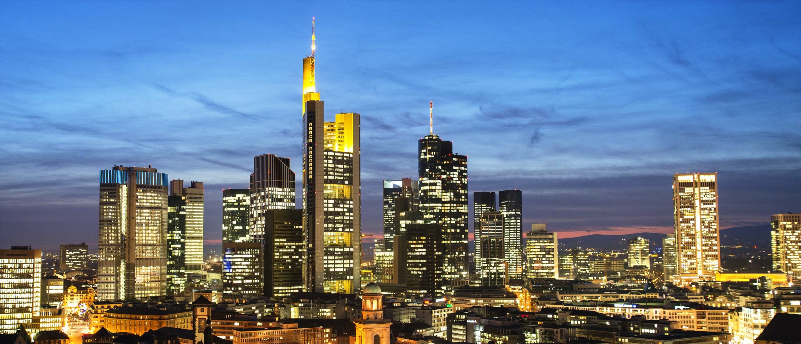 Die Skyline von Frankfurt als Beispiel für erfolgreiche Immobilien-Projektentwicklung in Deutschland. Ein Wald von leuchtenden Hochhaus-Immobilien im nächtlichen Abendhimmel. Unternehmen für Immobilien-Entwicklung erwerben und entwickeln Immobilien.